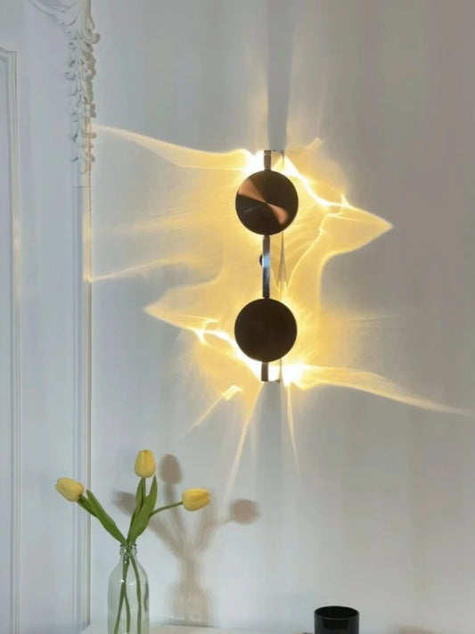 A modern wall sconce with two black circular discs mounted vertically on a wall, casting dramatic light patterns.