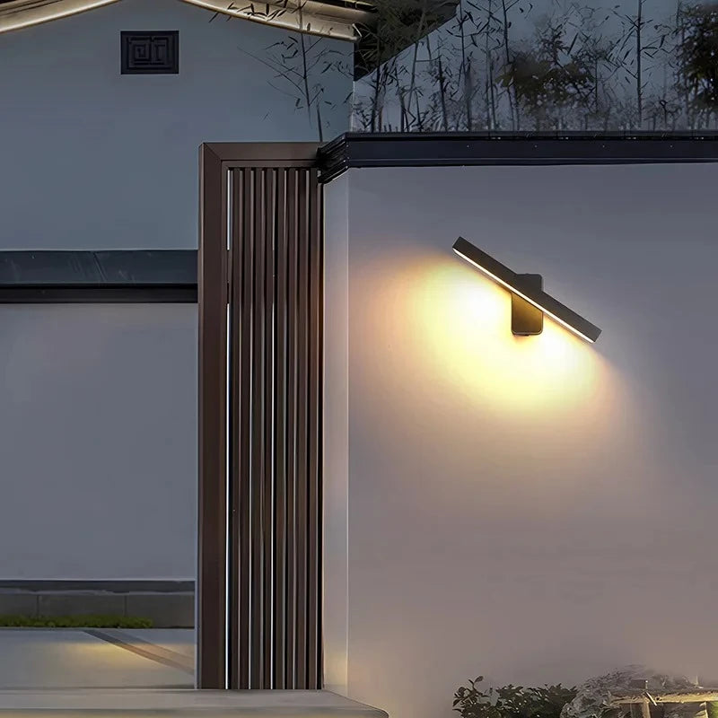 Black rotatable LED wall sconce mounted on an exterior wall, illuminating a pathway at night.
