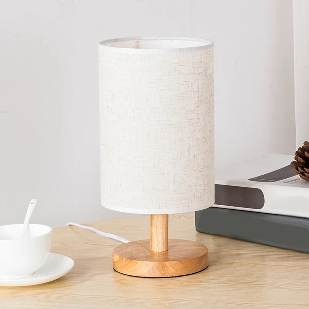 A plain white linen drum shade table lamp with a natural wood base, positioned on a wooden table with a white cup and saucer nearby.