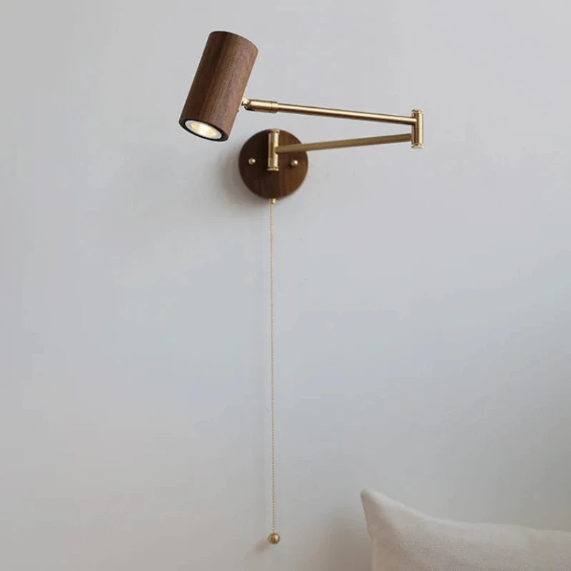 A modern wall sconce with a dark wood cylinder lamp head and a brass swing arm mounted on a round wooden base against a white wall, with a brass pull chain hanging down.