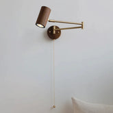 A modern wall sconce with a dark wood cylinder lamp head and a brass swing arm mounted on a round wooden base against a white wall, with a brass pull chain hanging down.
