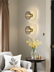 Two modern wall sconces with smoky gray glass diffusers mounted on a neutral-toned wall in a living room setting, casting circular light patterns.