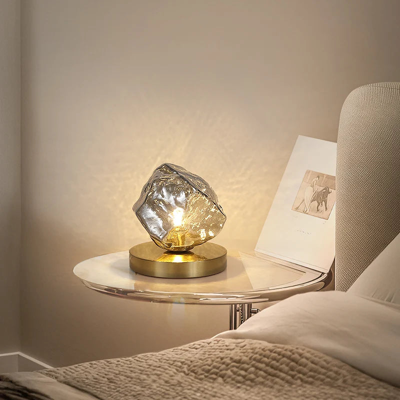 Glass table lamp on a nightstand next to a bed with soft lighting