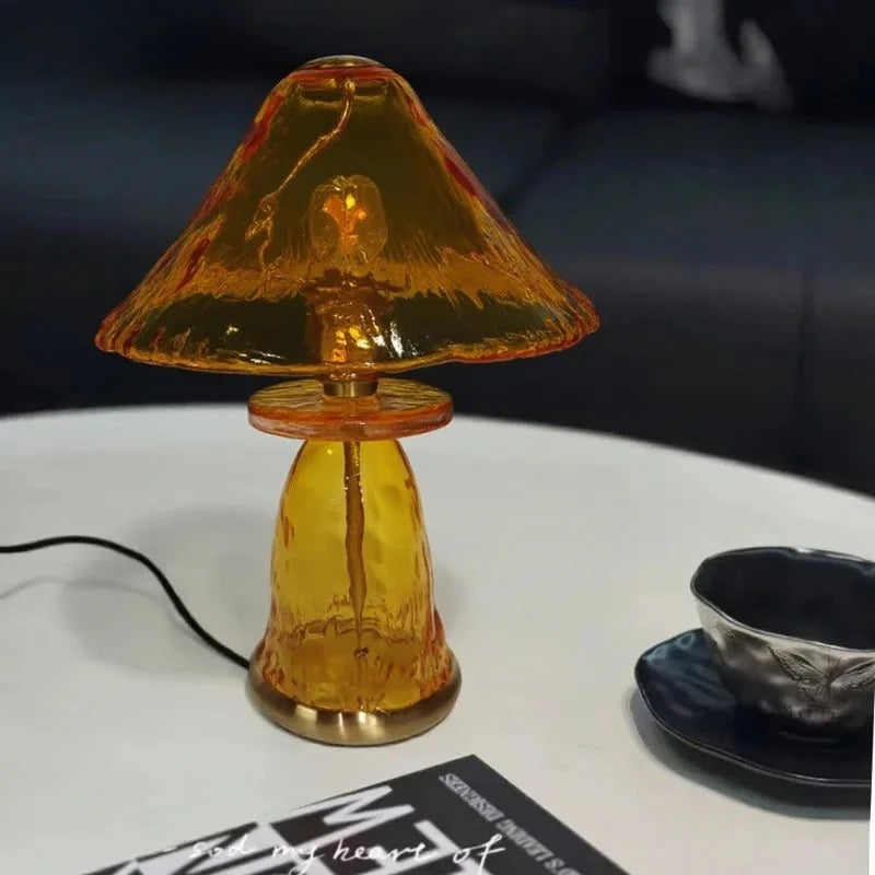 A bright amber glass mushroom table lamp sits on a white surface next to a modern dark bowl.