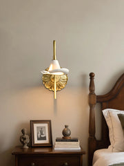 A gold floral ceramic wall sconce with a curved arm and white shade, mounted on a beige wall next to a wooden bed frame and nightstand.
