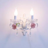A white wall sconce with two flame-tip bulbs, adorned with pink ceramic roses and clear crystal droplets hanging from a clear glass bowl-shaped holder.