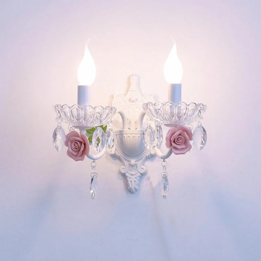 A white wall sconce with two flame-tip bulbs, adorned with pink ceramic roses and clear crystal droplets hanging from a clear glass bowl-shaped holder.