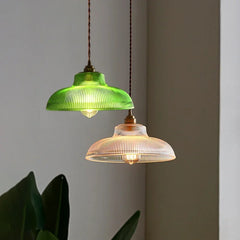 Two fluted glass dome pendant lights, one green and one clear with a polished copper finish, hanging in a room with a large plant.