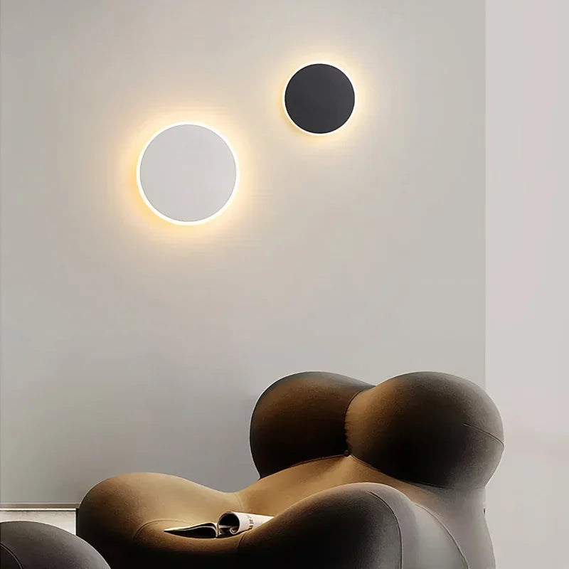 A white and a black circular wall sconce with warm halo effects mounted on a light-colored wall above a brown armchair.