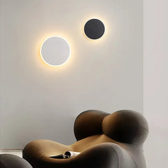 A white and a black circular wall sconce with warm halo effects mounted on a light-colored wall above a brown armchair.