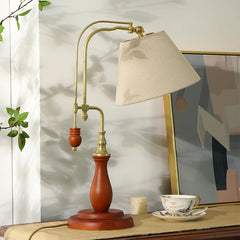 Table lamp with beige shade on a wooden surface, with a plant and abstract painting in the background.