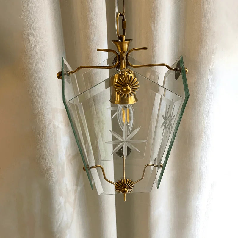 A clear glass pendant light with etched starburst patterns and brass accents, hanging against a curtain.