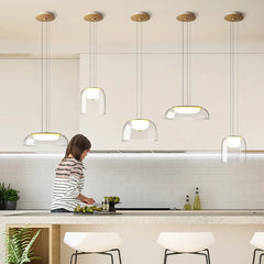 A woman in a kitchen preparing food, with five clear glass dome pendant lights with gold accents installed above the counter.