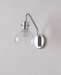 Wall-mounted lamp with clear glass shade and chrome finish on a light gray wall.