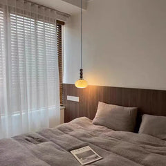 A wood bead and yellow persimmon-shaped globe pendant light hanging above a bed with white blinds in the background.
