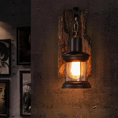 Close-up of a black iron lantern wall sconce with a distressed wood backplate mounted on a textured gray wall, with framed art on the left.