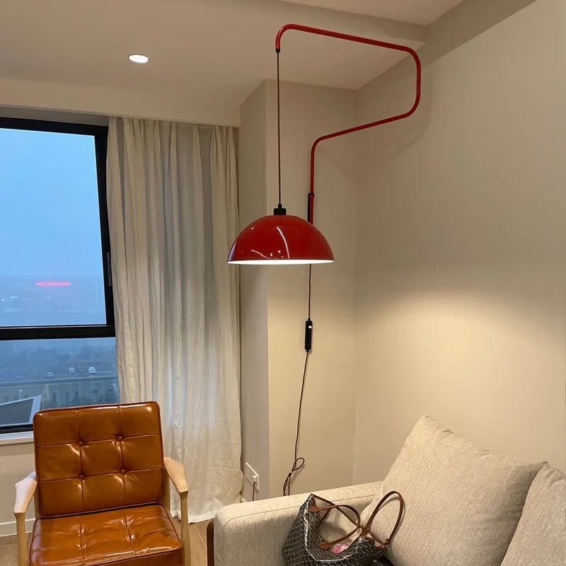 Red folding retractable wall lamp with a dome shade illuminating a corner of a living room with a large window and a leather armchair.