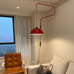 Red folding retractable wall lamp with a dome shade illuminating a corner of a living room with a large window and a leather armchair.