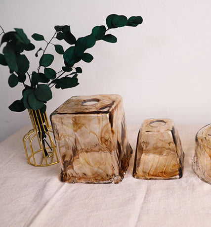 Set of four brown glass candle holders on a white surface with a plant in the background.