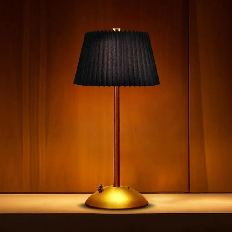 A black pleated fabric shade table lamp with a gold base and stem, illuminated against a warm wooden backdrop.