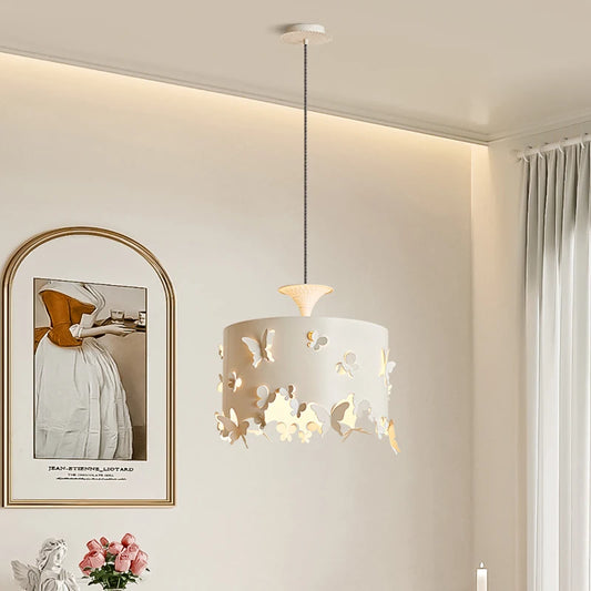 A cream-colored drum pendant light with intricate butterfly cutouts, hanging from a white ceiling. The light casts a warm glow through the cutouts.