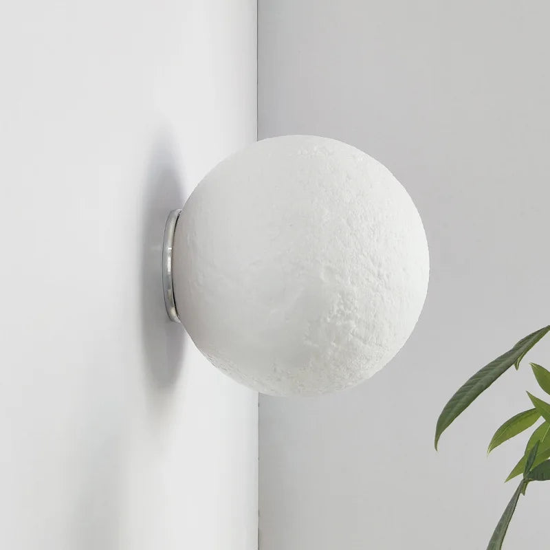 A white 3D printed moon shaped wall sconce with a textured surface is mounted on a white wall, casting a subtle shadow. A green leaf plant is in the lower right foreground.