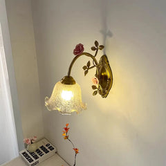 A gold floral wall sconce with a pink rose and fluted glass shade casts a warm light on a white wall, with a stack of books visible below.