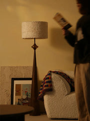 A dark grey floor lamp with a floral drum shade stands in a softly lit room next to a textured armchair.