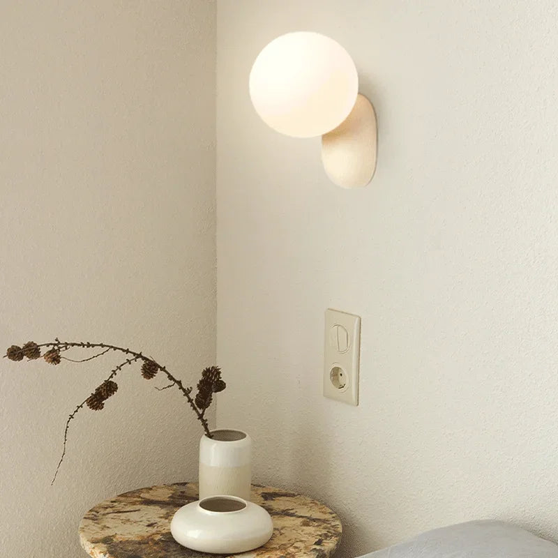 A minimalist wall sconce with a beige globe shade mounted on a light gray wall, with a decorative vase on a side table below.