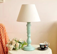 Light blue table lamp with white shade on a surface with flowers and a cup.