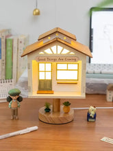 White wooden tree house shaped table lamp with a green door, windows, and a sign that reads 'Good Things Are Coming', illuminated by warm LED lights.