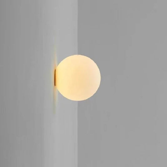 A white frosted glass sphere wall sconce mounted on a light gray corner wall, emitting a soft warm light.