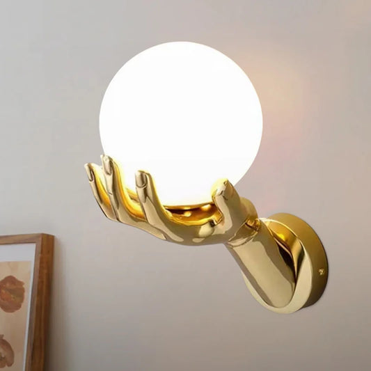 Gold hand wall sconce holding a white globe light against a light-colored wall.