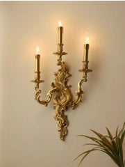 A close-up of a gold ornate wall sconce featuring three candle-shaped bulbs, highlighting its intricate details.