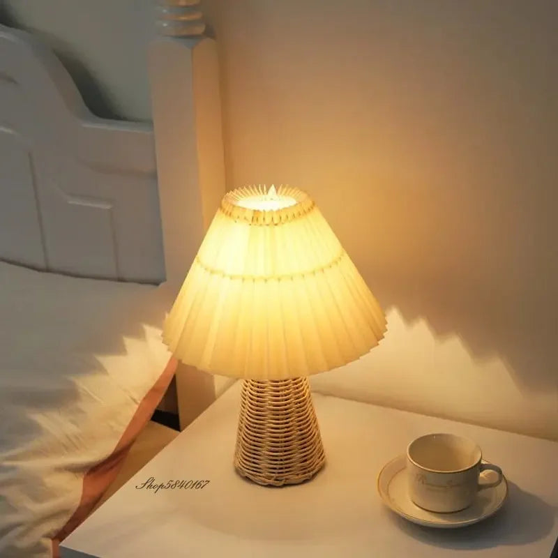 Table lamp with a warm white pleated fabric shade and a woven rattan base, lit on a bedside table next to a cup and saucer.