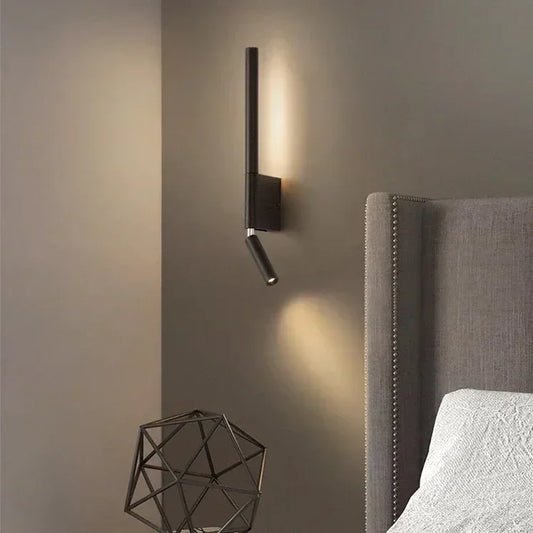 A modern black wall sconce with an adjustable arm, mounted on a grey wall next to a bed.