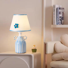 Blue and white striped ceramic table lamp with a scalloped white fabric shade featuring a blue flower accent, placed on a white nightstand next to a shelf with books.