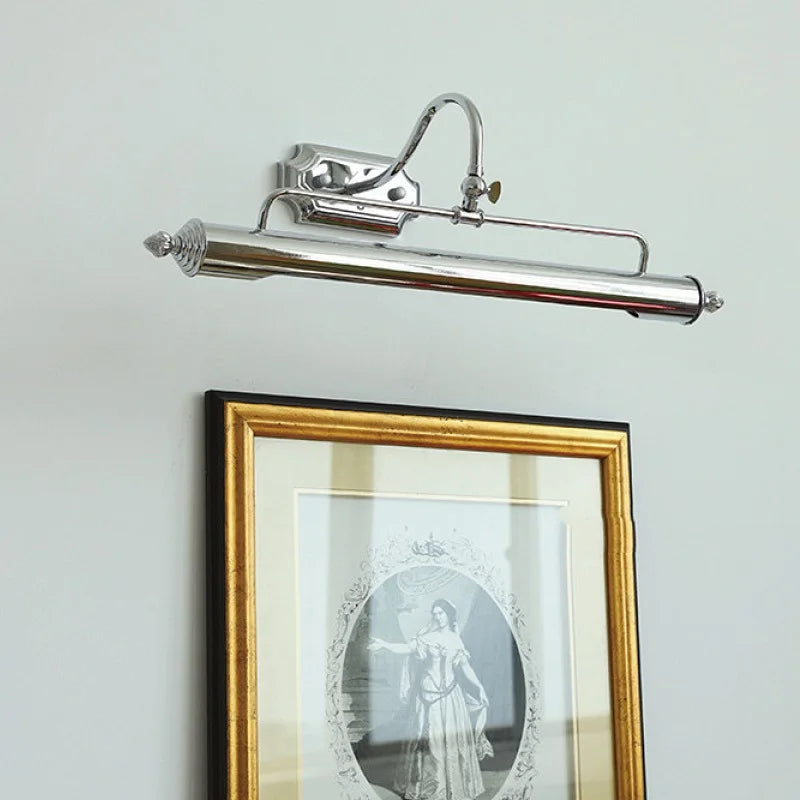 A silver-colored linear wall sconce with an adjustable arm and shade is mounted on a white wall above a framed picture.