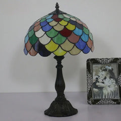 A Tiffany-style table lamp with a multicolored stained glass dome shade and a dark iron base, positioned on a surface.
