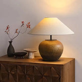 A ceramic pot-shaped table lamp with a conical fabric lampshade rests on a wooden dresser, with decorative items nearby.