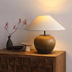 A ceramic pot-shaped table lamp with a conical fabric lampshade rests on a wooden dresser, with decorative items nearby.