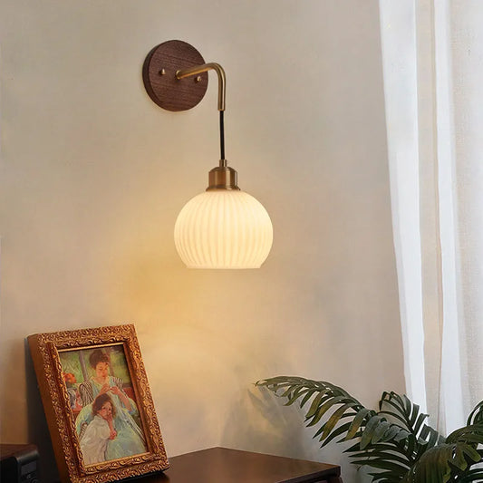 Wall sconce with a round walnut wood backplate, a curved brass arm, and a ribbed frosted glass globe shade, illuminated against a wall.