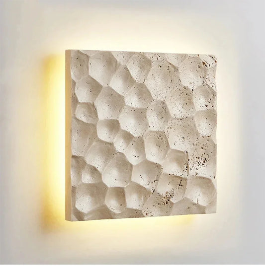 A square wall sconce with a textured, honeycomb-like pattern in a light stone finish, emitting a warm yellow glow from behind.
