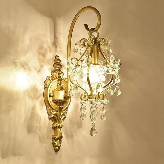 A close-up, angled view of a gold wall sconce with cascading clear crystals illuminated against a neutral wall, casting a warm glow and patterned light.