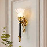 A gold wall sconce with a flared clear glass shade and a dark conical body, mounted on a white door.