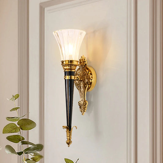 A gold wall sconce with a flared clear glass shade and a dark conical body, mounted on a white door.