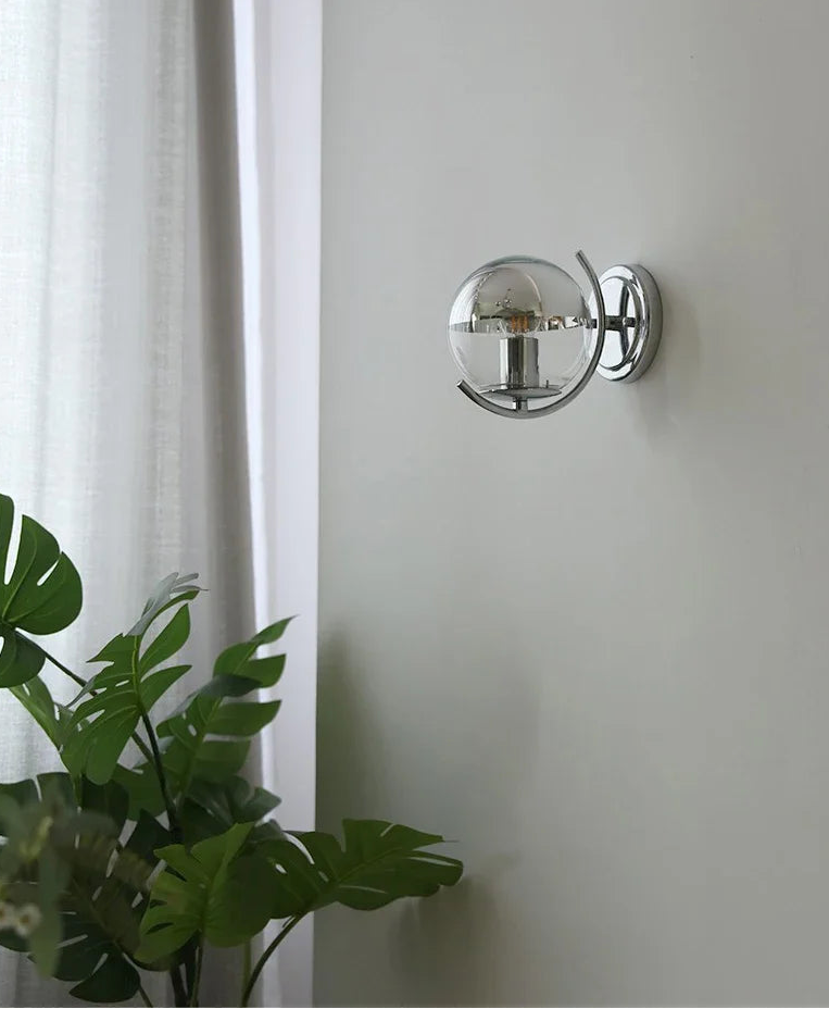 Wall-mounted spherical light fixture on a white wall with a plant in the foreground.