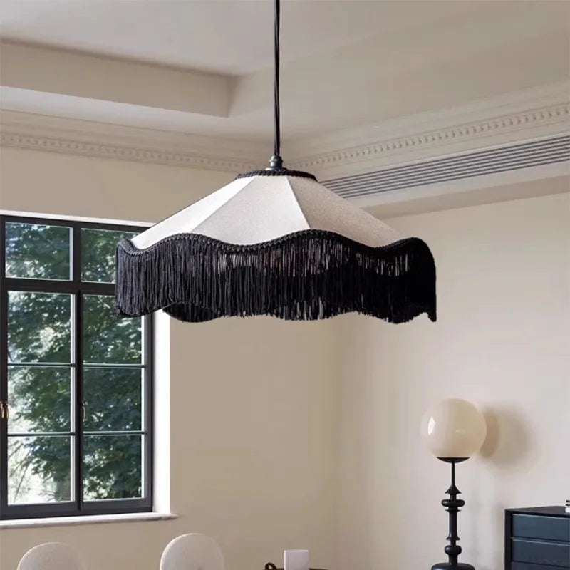 Black and white fabric pendant light with long black tassels hanging from a scalloped edge, shown in a room with a large window.