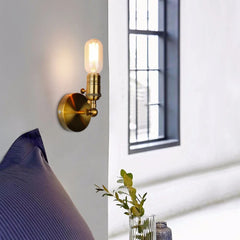 Close-up of a gold industrial-style wall sconce with an adjustable folding arm and exposed Edison-style bulb, mounted on a white wall next to a blue pillow.