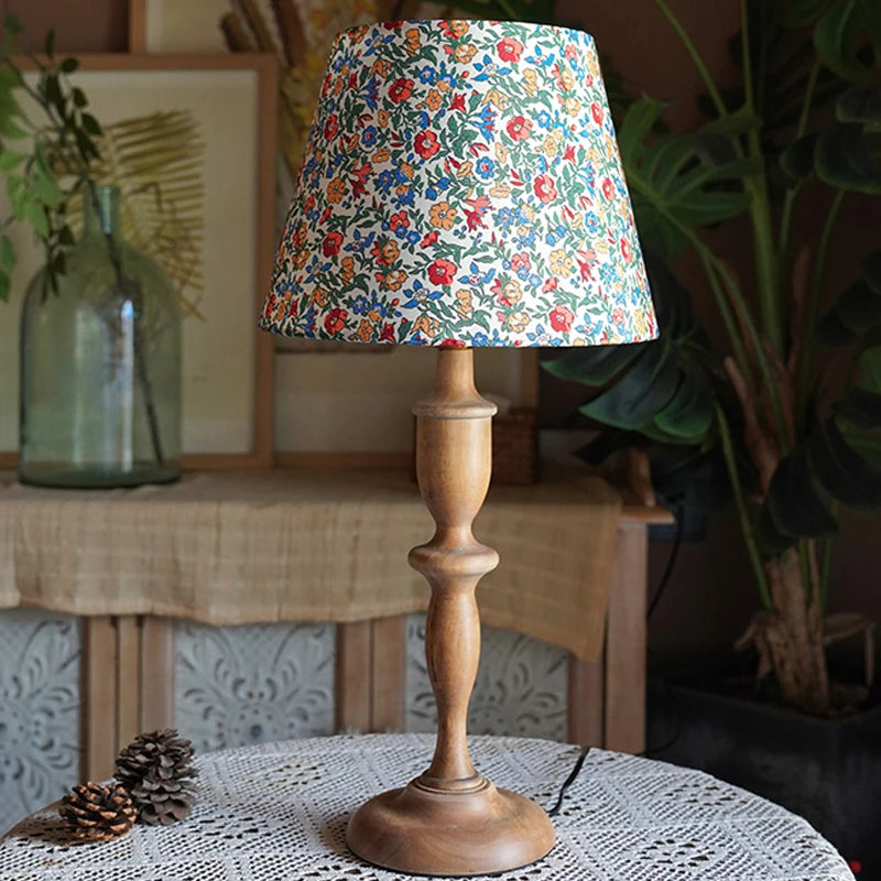 Decorative lamp with floral shade on a wooden base, placed on a table with plants in the background.